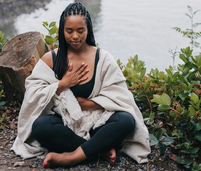 A young woman of color is contently sitting cross legged near a pond.  She has one hand on her chest and the other on her belly